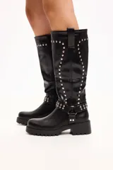 Bota de caña media en cuero sintético negro, con suela track, cierre lateral y detalle de tachas metálicas en la parte superior y en una tira decorativa sobre el tobillo.