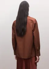 Camisa de algodón color marrón, de diseño cruzado estilo sobre con lazo lateral para anudar, cuello camisero, manga larga y calce amplio.