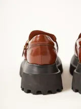 Mocasines marrones de plataforma con suela track negra y capellada de cuero. Presentan una tira en el empeine con hebilla dorada cuadrada y pespuntes visibles en contraste.