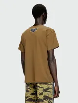 Remera de manga corta color marrón, con cuello redondo y un pequeño parche estampado con diseño abstracto en el pecho.