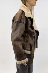 Campera oversize de efecto cuero color marrón con interior, solapas, cuello y puños de corderito sintético en tono tostado. Cuenta con detalles de tiras ajustables con hebilla en el cuello, mangas y ruedo, y cierre frontal cruzado.