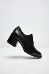 Zapato de cuero negro con tacón grueso y suela dentada. Presenta una combinación de texturas, con detalles de cuero grabado en el talón y el lateral.