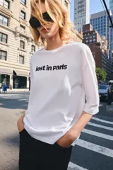 Remera oversize blanca de algodón con cuello redondo, mangas amplias por debajo del codo y estampado frontal con el texto "lost in Paris" en color negro.