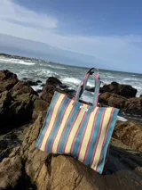 Bolso tipo tote grande con estampado a rayas verticales en tonos celeste, blanco y bordó. Tiene doble asa corta de mano y cierre superior.