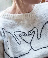 Sweater artesanal de lana merino en color natural, con diseño tejido de jacquard en la parte inferior y bordado a mano de dos cisnes enfrentados formando un corazón en el pecho. Presenta cuello redondo, puños y ruedo en tono verde contrastante.