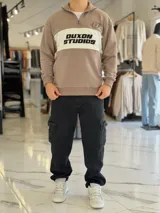 Hoodie de corte clásico con medio cierre y cuello alto, en color marrón con una franja horizontal blanca en el pecho que incluye el logo estampado en negro. Presenta además un pequeño bordado en el hombro izquierdo y puños y cintura elastizados de rib.