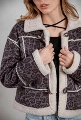Campera de gamuza con estampado animal print en tonos grises, forrada con corderito sintético en color crema. Presenta cierre frontal con botones tipo toggle y detalles de costuras visibles.