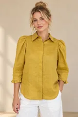 Camisa de lino color blanca, con cuello clásico, mangas tres cuartos abullonadas en los hombros y terminación con volados en los puños, cierre frontal con botones ocultos.