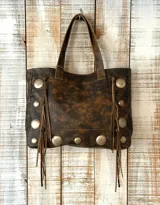 Cartera tipo tote de cuero con efecto desgastado, decorada con tachas metálicas circulares y flecos laterales. Cuenta con doble asa de hombro.
