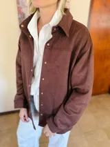 Sobrecamisa de gamuza con cuello clásico, cierre frontal con botones metálicos y mangas largas con puños abotonados. Presenta un diseño estructurado con corte recto y largo a la cadera.