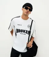 Remera de manga corta en tejido sintético color blanco, con cuello redondo y diseño deportivo. Presenta estampado frontal con tipografía gótica y detalles gráficos, además de franjas laterales negras y paneles con perforaciones para mayor ventilación.