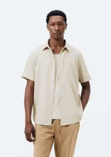 Camisa de manga corta color beige, confeccionada en tejido de lino, con cuello camisero y cierre frontal de botones.