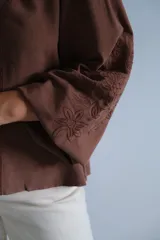 Blusa color chocolate con cuello en V, cierre frontal con botones y mangas largas abullonadas con puños elásticos. Presenta detalles de bordado lineal a lo largo de la abertura frontal.