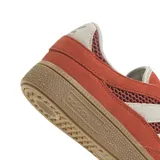 Zapatilla Adidas Handball Spezial, color naranja quemado con tres franjas laterales en color crema. Presenta paneles laterales de malla y puntera de gamuza. La suela es de goma color gum (marrón claro).
