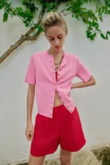 Chaqueta de punto de manga corta en color rosado, con cuello redondo y cierre frontal de botones dorados.