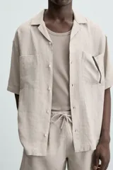 Camisa de lino de calce relajado en color beige, con cuello bowling y manga corta. Cuenta con dos bolsillos de parche en el pecho, aberturas laterales en el bajo y cierre frontal con botones.