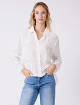 Camisa de gasa ligera y traslúcida, color blanco, con intrincados bordados búlgaros y motivos botánicos distribuidos en toda la prenda. Presenta una guarda bordada que recorre la línea de botones, el ruedo y los puños. Tiene cuello con solapa y abertura central con botones. La silueta es amplia y relajada, con mangas largas y un canesú en la espalda para una caída impecable.