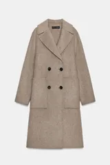 Abrigo largo color beige, confeccionado con lana, con cuello solapa, manga larga y bolsillos delanteros de plastrón. Cierre frontal cruzado con botones negros.