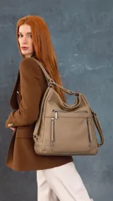 Cartera mochila versátil de cuero sintético con textura granulada, color beige. Cuenta con múltiples bolsillos frontales con cierre, herrajes metálicos y correas ajustables que permiten llevarla tanto al hombro como mochila.