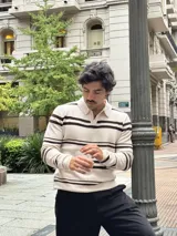 Buzo tejido de punto con diseño a rayas horizontales en color beige y marrón. Presenta cuello tipo polo con cierre frontal parcial y puños acanalados.