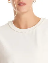 Blusa de crepe color blanco, con cuello redondo y mangas cortas. Presenta apliques de perlas decorativas en el borde del cuello y en los extremos de las mangas.