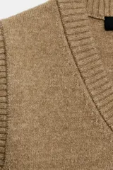 Chaleco de punto en color verde oliva, con escote en pico y sisas amplias. Presenta terminaciones acanaladas en el cuello, las sisas y el ruedo.