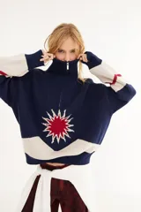 Sweater de punto de algodón con cuello polo y cierre frontal. Presenta un diseño con intarsia de sol y montañas en tonos azul, blanco y rojo, con detalle de parche en la manga.