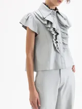 Camisa de cuero ovino color gris claro, con cuello camisero, volados centrales y mangas cortas con volumen generado por frunces en la copa. Cierre frontal con broches forrados.