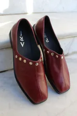 Zapato de cuero vacuno color bordó, con diseño de punta cuadrada y detalle de tachas metálicas planas en el borde superior. Presenta un taco geométrico bajo y costura central en el empeine.