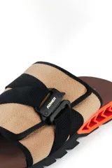 Chinelas Kenner modelo Rakka Slide Pro, con capellada de tela color beige y ajuste mediante hebilla técnica negra. Presenta una base ergonómica marrón con estructura lateral geométrica en color naranja y suela negra.