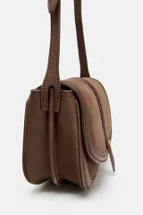 Bolso bandolera de diseño estructurado en color marrón, confeccionado en una combinación de cuero y gamuza. Presenta una solapa frontal con cierre imantado, detalle de tira central con remache metálico y correa larga ajustable para llevar al hombro o cruzado.
