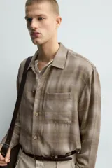 Camisa relaxed fit de manga larga con cuello solapa, confeccionada en tejido con mezcla de viscosa. Presenta un bolsillo plastrón en el pecho, bajo con aberturas laterales y cierre frontal de botonadura. El diseño es a cuadros en tonos beige y marrón.