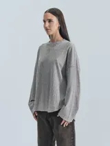 Sweater gris de tejido acanalado, manga larga y cuello redondo. Presenta un corte holgado con bajo redondeado y un pequeño parche negro con logo en la manga izquierda.