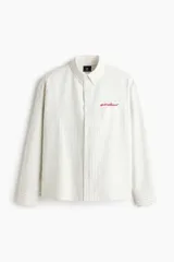 Camisa de corte holgado en mezcla de algodón Oxford, con diseño a rayas, cuello americano y cierre frontal de botones. Cuenta con un bolsillo superior en el pecho con texto bordado en rojo, mangas largas con puños abotonados y bajo recto.