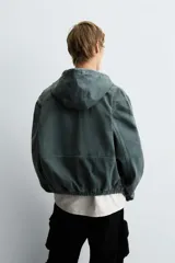 Cazadora negra de algodón con efecto lavado, corte relajado y capucha ajustable. Presenta cierre frontal de cremallera, bolsillos frontales tipo canguro y elásticos en puños y bajo.