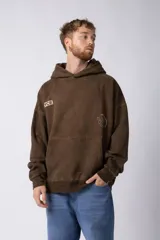 Buzo tipo canguro con capucha, de corte oversize y confeccionado en frisa rústica color marrón. Presenta estampado con texto en el pecho derecho y logo gráfico en el pecho izquierdo.