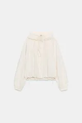 Chaqueta corta de nylon color beige, con cuello subido, capucha y cierre frontal con cremallera. Presenta mangas largas con puños elásticos, bolsillos laterales y detalles de cordones ajustables con lazadas en el pecho y el bajo.
