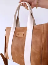 Cartera tipo tote de cuero color camel con detalles de bandas verticales en color crema. Cuenta con doble asa de mano, correa larga ajustable y extraíble, y cierre superior. Incluye etiqueta de cuero con logo grabado en el frente.