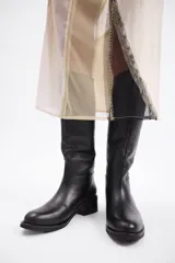 Bota de caña alta confeccionada en cuero vacuno, con taco cuadrado bajo y punta redondeada.