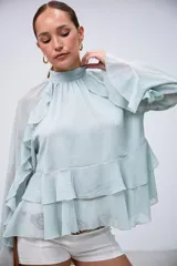 Blusa verde menta de gasa, con cuello alto fruncido, mangas largas amplias y detalles de volados en cascada en el frente y las mangas.