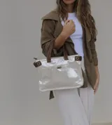 Bolso de mano tipo tote, confeccionado en cuero metalizado color plateado con detalles en cuero marrón en las asas y laterales. Cuenta con un bolsillo frontal con cierre giratorio y herrajes metálicos.