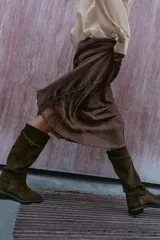 Bota de caña alta estilo hípico confeccionada en serraje color marrón oscuro. Presenta punta redonda, suela plana con pequeño tacón y detalle de tira con hebilla metálica en la parte superior de la caña.