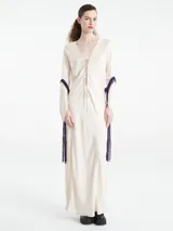 Vestido largo tipo kaftan color crema, confeccionado en una mezcla de cupro y rayón. Presenta un escote en V profundo con detalle de cordones cruzados y mangas largas adornadas con flecos largos en color violeta.