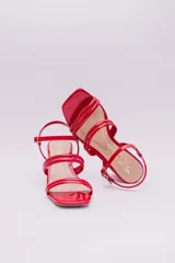 Sandalias de tacón cuadrado medio, confeccionadas en charol rojo. Presentan un diseño de tiras tubulares sobre el empeine y cierre con pulsera ajustable al tobillo mediante hebilla metálica.