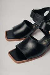 Sandalias de cuero negro con diseño minimalista y tacón ancho geométrico en tono madera. La capellada lisa aporta una estética depurada y moderna, mientras que la pulsera con hebilla metálica ajustable asegura un calce firme y confortable.