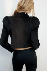 Chaqueta entallada negra de manga larga y cuello redondo, con apliques de tachas metálicas en los hombros, a lo largo de la abertura frontal y en los puños. Cuenta con cierre frontal con gancho metálico.