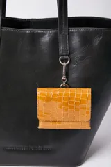 Cartera tipo shopper de cuero vacuno en color negro, con asas largas para hombro y estructura semirrígida. Incluye un pequeño monedero colgante desmontable de cuero texturizado en color mostaza en la parte frontal.