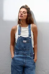 Jumpsuit de jean azul clásico con diseño de jardinera, tirantes ajustables con hebillas metálicas, bolsillo frontal con etiqueta de marca y bolsillos laterales.