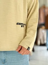 Sweater de punto liviano color beige, con textura marcada, cuello redondo y mangas largas de corte relajado, con pequeño estampado tipográfico negro en el lateral inferior.