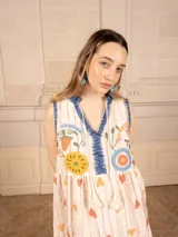 Vestido largo color crema con estampado de flores y corazones en tonos azules, naranjas y amarillos. Tiene cuello en V con ribete azul y corte imperio con falda escalonada.